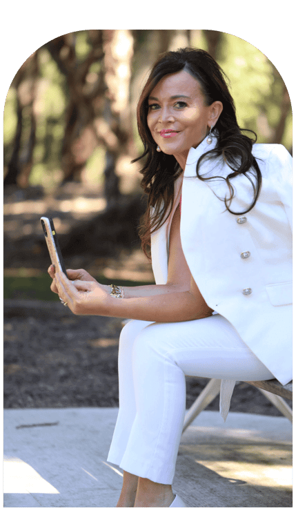 Terri sitting on a bench with her phone wearing a white jacket looking at the camera